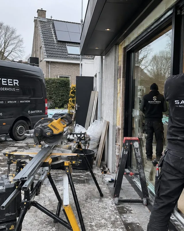 Twee medewerkers van SANTEER plaatsen glas in een schuifpui, met de bedrijfsbussen en een zaagtafel ervoor